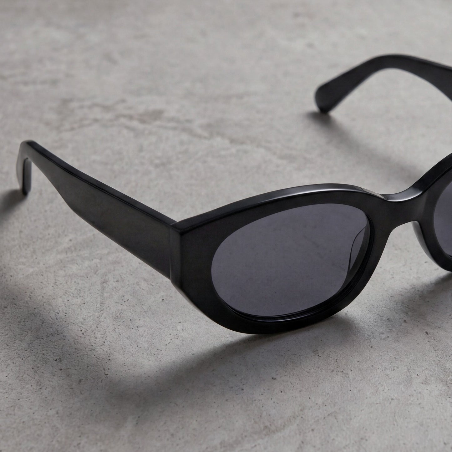 Close-up of black oval acetate sunglasses resting on textured stone surface in soft directional light.