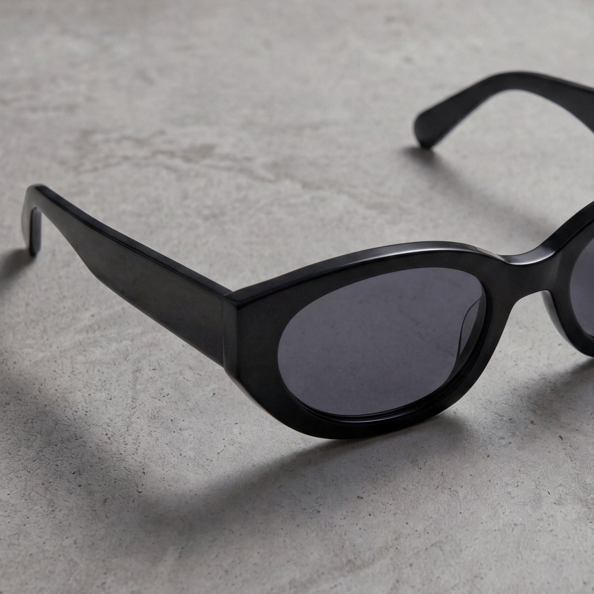 Close-up of black oval acetate sunglasses resting on textured stone surface in soft directional light.
